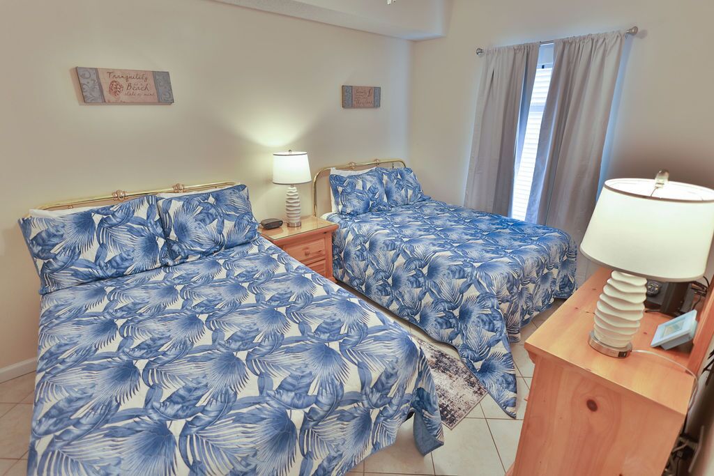A room with two beds, each with a blue floral bedspread, a bedside table with a lamp, and a small wooden nightstand.