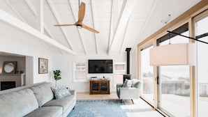 A spacious living room with a large gray sofa, a blue patterned rug, a wooden ceiling fan, and a large window with a view of the outdoors.
