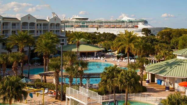 Un grand navire de croisière a accosté dans une station balnéaire dotée de plusieurs piscines, de palmiers et d’un bâtiment d’hôtel.