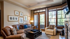 A living room with a brown leather sofa, a white armchair, a coffee table, and a fireplace. There are framed pictures on the wall and a large window with a view of trees.