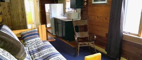 Un acogedor interior de cabaña con piso de madera, un sofá con cojines estampados, una mesa de comedor con una silla, una zona de cocina con refrigerador y microondas, y una ventana con cortinas.