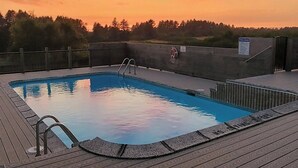 A swimming pool with a wooden deck, surrounded by trees, under a sunset sky. - Private Executive Home with Pool, Pickleball Court, and Sauna. (Gearhart)
