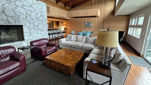 A spacious living room with a stone wall, wooden ceiling beams, a fireplace, leather sofas, a coffee table, and a dining area with chairs and a table. - Private Executive Home with Pool, Pickleball Court, and Sauna. (Gearhart)