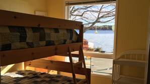 A bunk bed with a quilt, a wooden ladder, a white crib, and a view of a lake through a window.