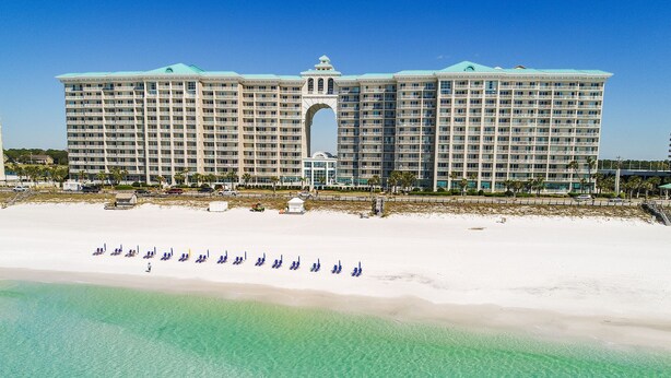 Un grand hôtel doté d’une arche centrale, une plage bordée de rangées de parasols et une eau turquoise et cristalline.