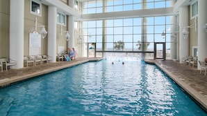 Une piscine intérieure offrant une vue dégagée sur le ciel grâce à de grandes fenêtres, entourée de colonnes et de fauteuils blancs.