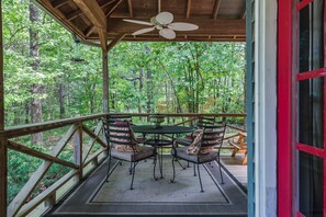A covered outdoor seating area with a table and chairs, surrounded by a wooden deck and a forest view.