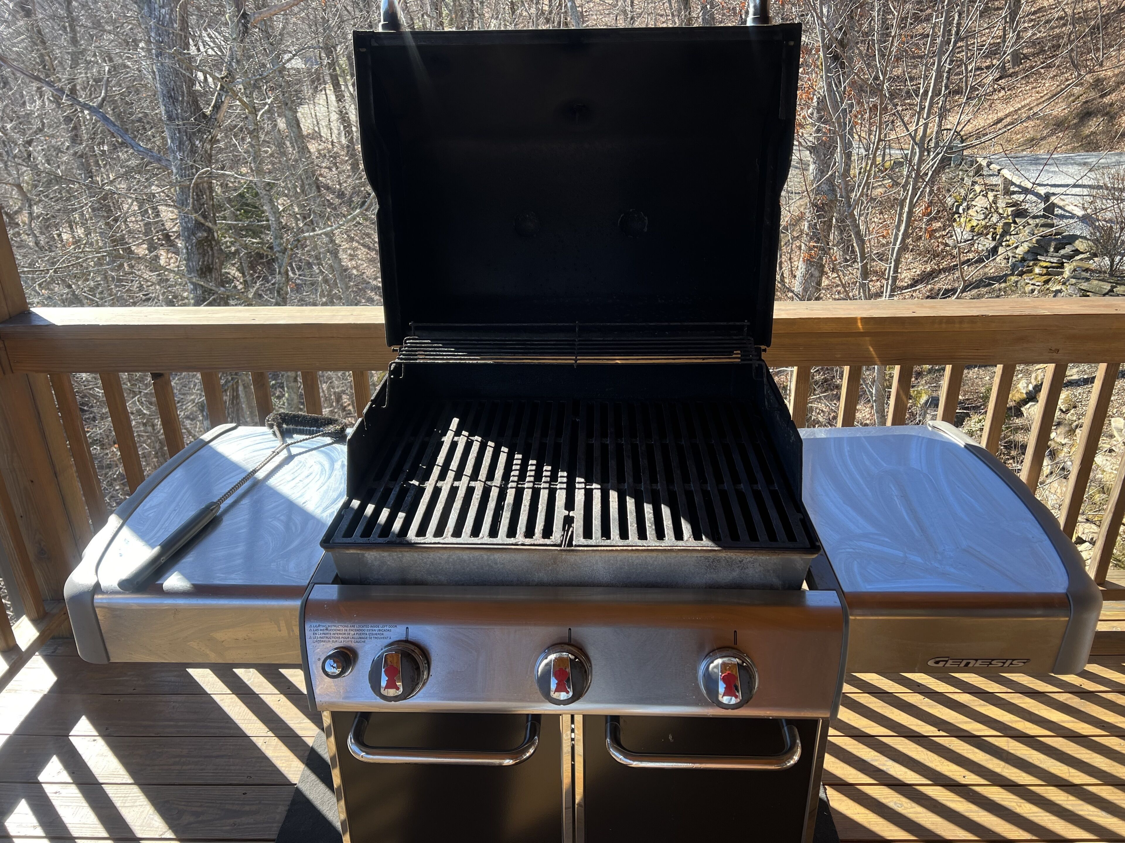 Un barbecue portable de la marque Genesis, placé sur une terrasse en bois, avec vue sur des arbres et un mur de pierres en arrière-plan.