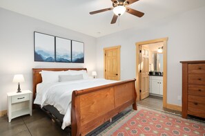 A bedroom with a wooden bed, a nightstand with a lamp, a ceiling fan, and a view into the kitchen through an open door. - Charming 5-star house near downtown Asheville & Biltmore, fire pit, dog friendly (Asheville)