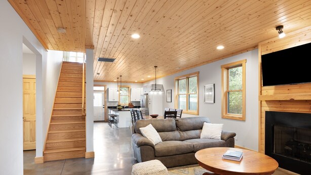 A spacious living area with a wooden staircase, a fireplace, a sofa, ottomans, and a dining area in the background.