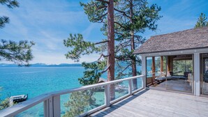 A wooden deck with glass railings overlooking a body of water, a building with large windows, and a mountainous backdrop. - Peaceful Lakefront Home with Gorgeous Lakeviews and Hottub, Lake St. Lakefront by Tahoe Time VR (Kings Beach)