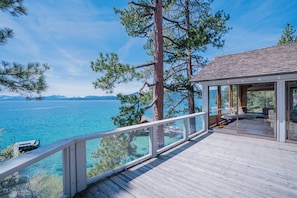 A wooden deck with glass railings overlooking a body of water, a building with large windows, and a mountainous backdrop.