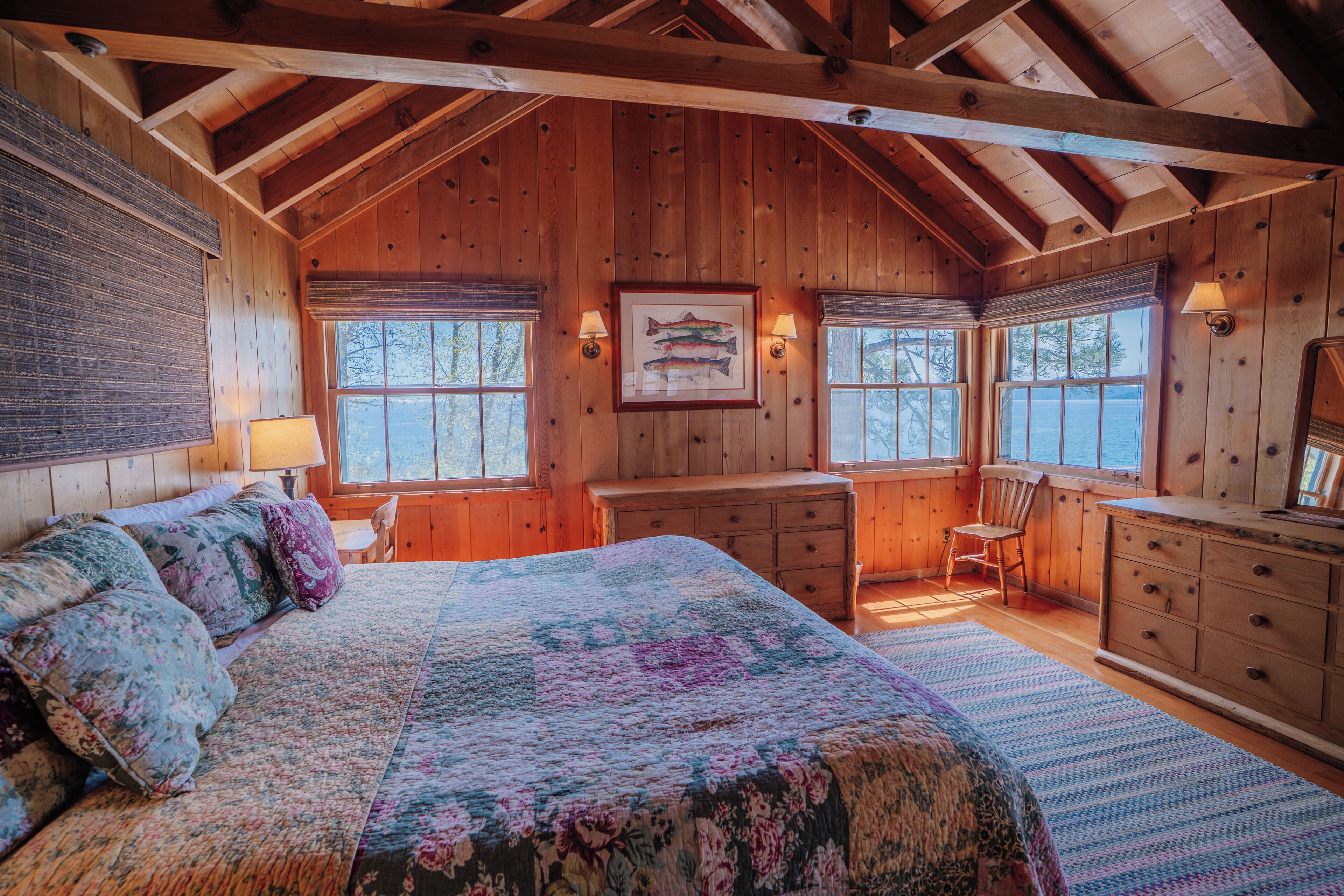 A wooden cabin bedroom with a bed, dresser, chair, and windows.