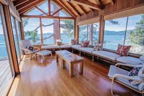 A wooden interior with a large window overlooking a body of water, a wooden bench, and a cushioned seating area.