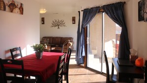 A dining area with a red tablecloth, chairs, a potted plant, and a sofa in the background.