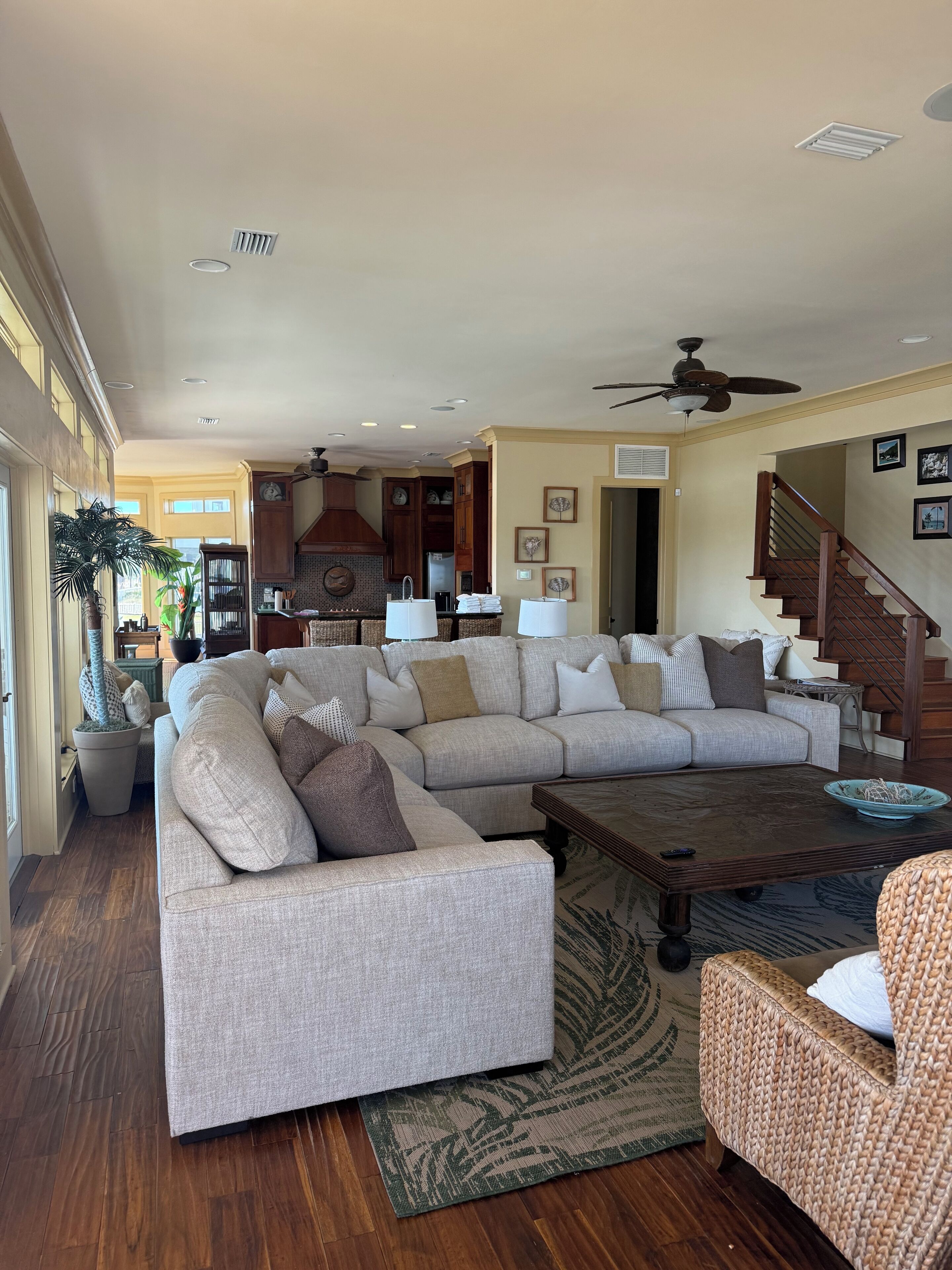 A spacious living room with a large sectional sofa, a coffee table, and a wicker chair. There is a kitchen area with wooden cabinets and a built-in oven. A staircase with wooden railings is visible in the background.