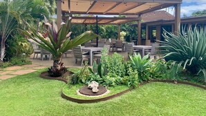 An outdoor patio area with tables and chairs, surrounded by tropical plants and a decorative stone feature. - A403 - Spacious 1 Bedroom, 2 King Bed, Garden and Partial Ocean Views (Lahaina)