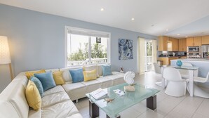 A modern living room with a glass coffee table, a white sofa with colorful cushions, and a view of the kitchen in the background. - Modern Beach House • Heated Pool • 3 Min Walk to Beach (South Padre Island)