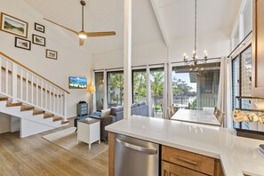 A modern kitchen with a central island, a dining area with a view, a staircase with wooden steps, and a living area with a sofa and a TV.