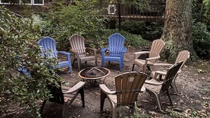 An outdoor seating area with Adirondack chairs and a fire pit, surrounded by trees and a house in the background. - Mountain House at the Beach! Dog Friendly, Short Walk to the Beach. (Villas)