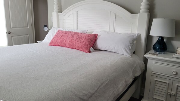 Een netjes opgemaakt bed met witte en roze kussens, een wit hoofdeinde, en een nachtkastje met een lamp en een klein voorwerp met de tekst 'BEACH'.