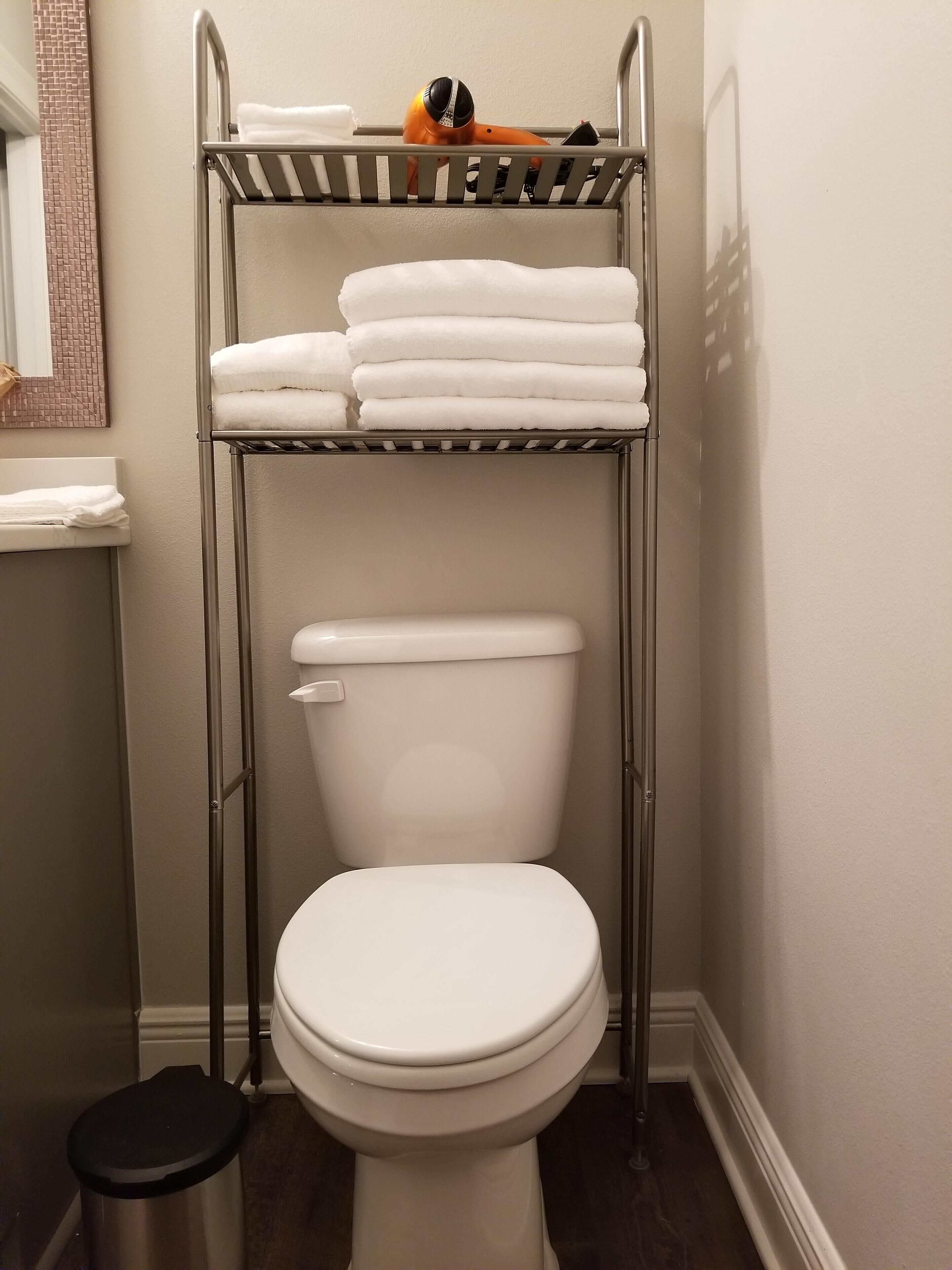A bathroom with a white toilet, a metal shelf with folded white towels, and a small orange object on the shelf.
