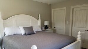 Ein ordentlich bezogenes Bett mit weißem Kopfteil und Fußteil, einer grauen Bettdecke und zwei Kissen. Ein Nachttisch mit Lampe und einer kleinen Dekoration. Die Wände des Zimmers sind beige und es gibt zwei weiße Türen.