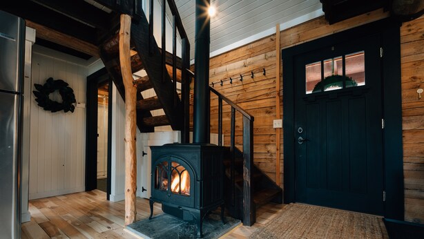 A cozy interior with a wood-burning stove, a staircase, and a dark door. - Secluded Rustic-Modern Log Cabin w/ Hot Tub, Private Creekside Escape (Ferryville)