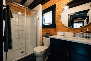 A bathroom with a glass shower enclosure, a toilet, a sink with a mirror, and wooden wall paneling. - Secluded Rustic-Modern Log Cabin w/ Hot Tub, Private Creekside Escape (Ferryville)