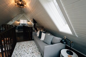 A cozy attic bedroom with a sloped ceiling, a gray sofa with pillows, a patterned rug, a small round table with a lamp, and a staircase leading to another room. - Secluded Rustic-Modern Log Cabin w/ Hot Tub, Private Creekside Escape (Ferryville)