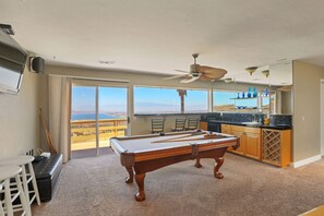 A pool table in a room with a large window, a ceiling fan, and a kitchen area. - Highest reviewed property in the area - best view of  your life! (Lake Mathews)