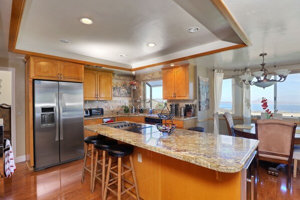 A modern kitchen with a large island, stainless steel refrigerator, wooden cabinets, and a view to the outside. - Highest reviewed property in the area - best view of  your life! (Lake Mathews)