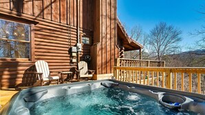 A wooden cabin with a hot tub on the deck, wooden chairs, and a view of the surrounding landscape.