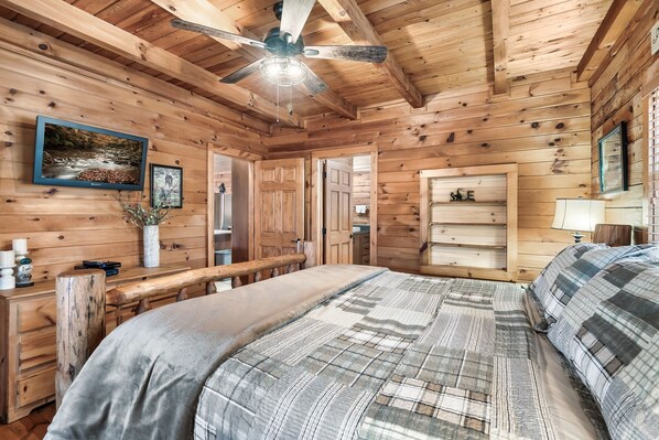 A wooden cabin bedroom with a bed, a desk, a ceiling fan, and a framed picture on the wall.