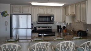 A kitchen with white cabinets, a refrigerator, a microwave, an oven, and a sink. There are chairs with blue cushions at the dining table. - Oceanfront Heaven in Exclusive West End, Ocean Isle Beach, NC  (Ocean Isle Beach)