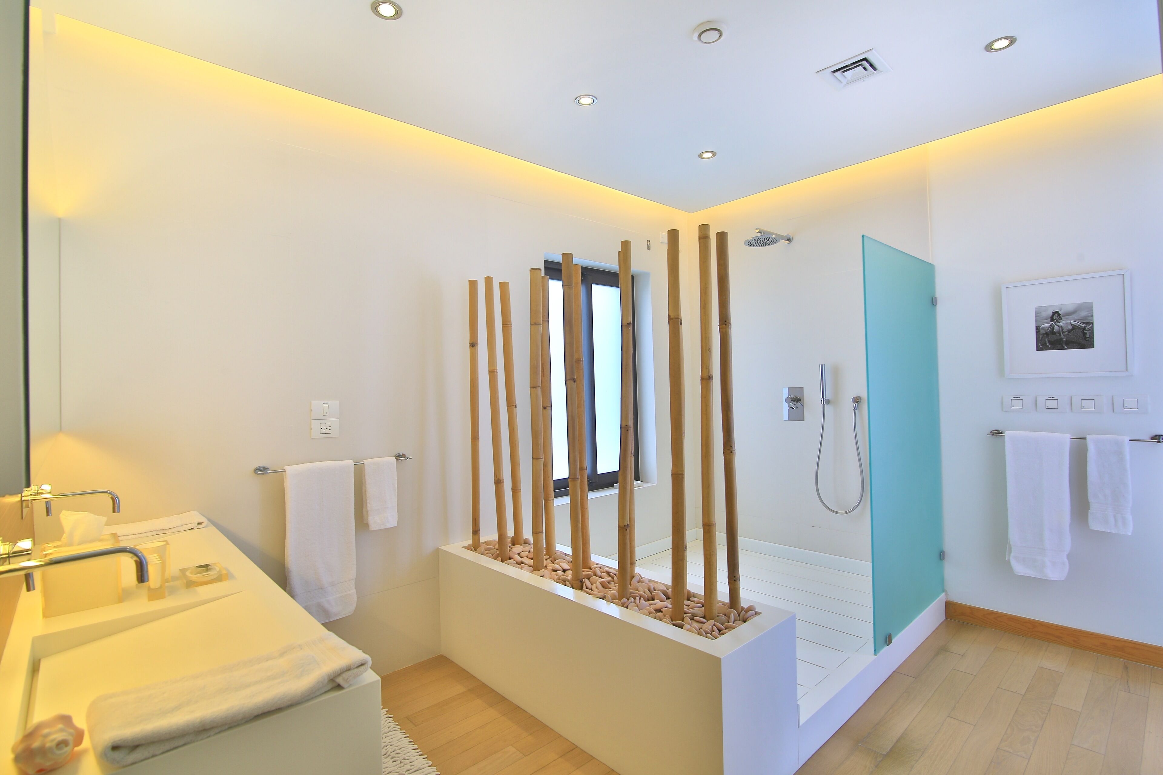 A modern bathroom with a white sink, mirror, and shower area. There are bamboo sticks in a planter, a white towel on the wall, and a picture on the wall.