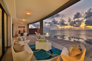 Un espace de détente extérieur moderne avec vue sur l’océan au coucher du soleil, comprenant un ensemble de canapés blancs, une table basse et une terrasse en bois.