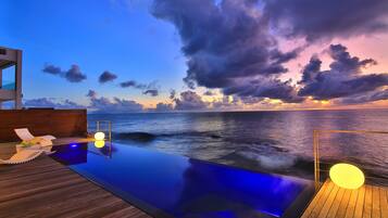 A wooden deck with a pool, lounge chairs, and a sunset view over the ocean.