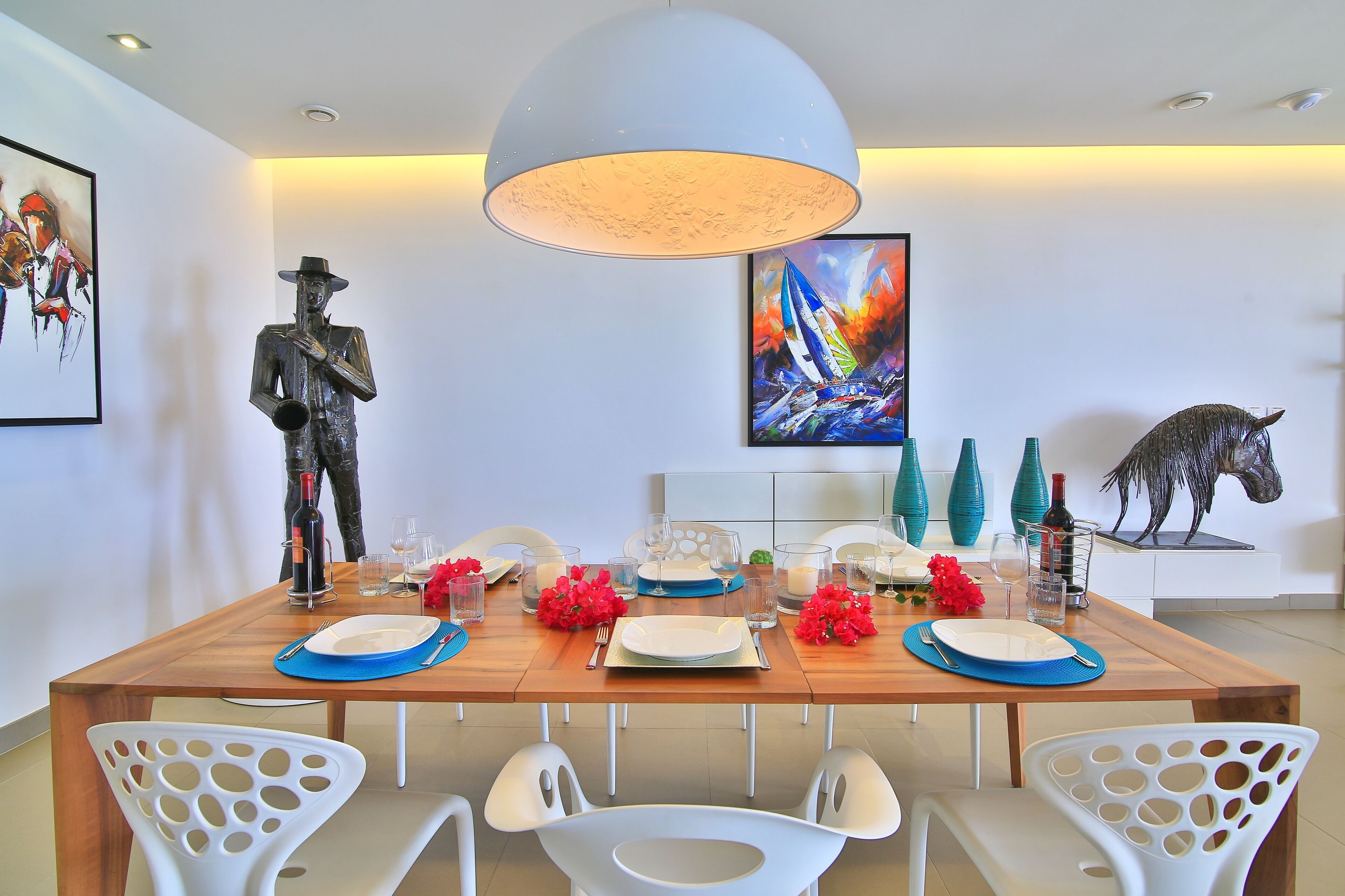 A dining area with a wooden table set for four, white chairs with cut-out backs, a large pendant light, and artwork on the walls.