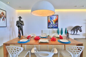 A dining area with a wooden table set for four, white chairs with cut-out backs, a large pendant light, and artwork on the walls. - Anchor House - Direct Oceanfront - Luxury & Modern Style (Cupecoy)