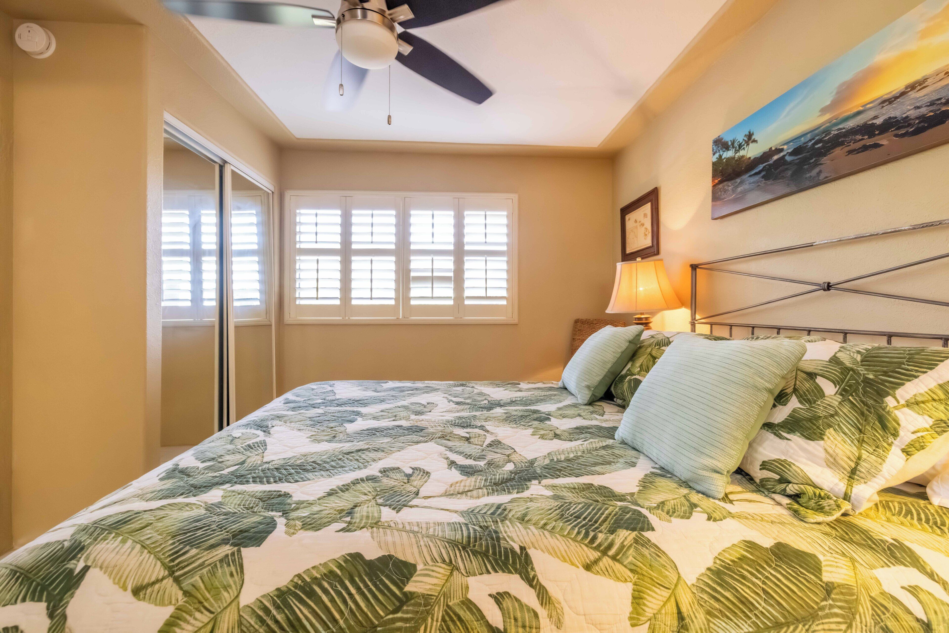 A bedroom with a bed, a ceiling fan, a window with blinds, a wall-mounted lamp, and a painting.