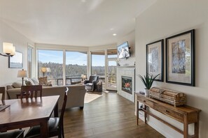 Relax in a spacious living area with a high ceiling and forest views, featuring a fireplace, dining table, sofa, and TV.