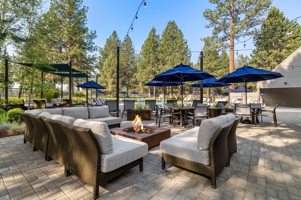 Relax in a serene outdoor space with wicker sofas, chairs, and fire feature, surrounded by tall trees and umbrellas.