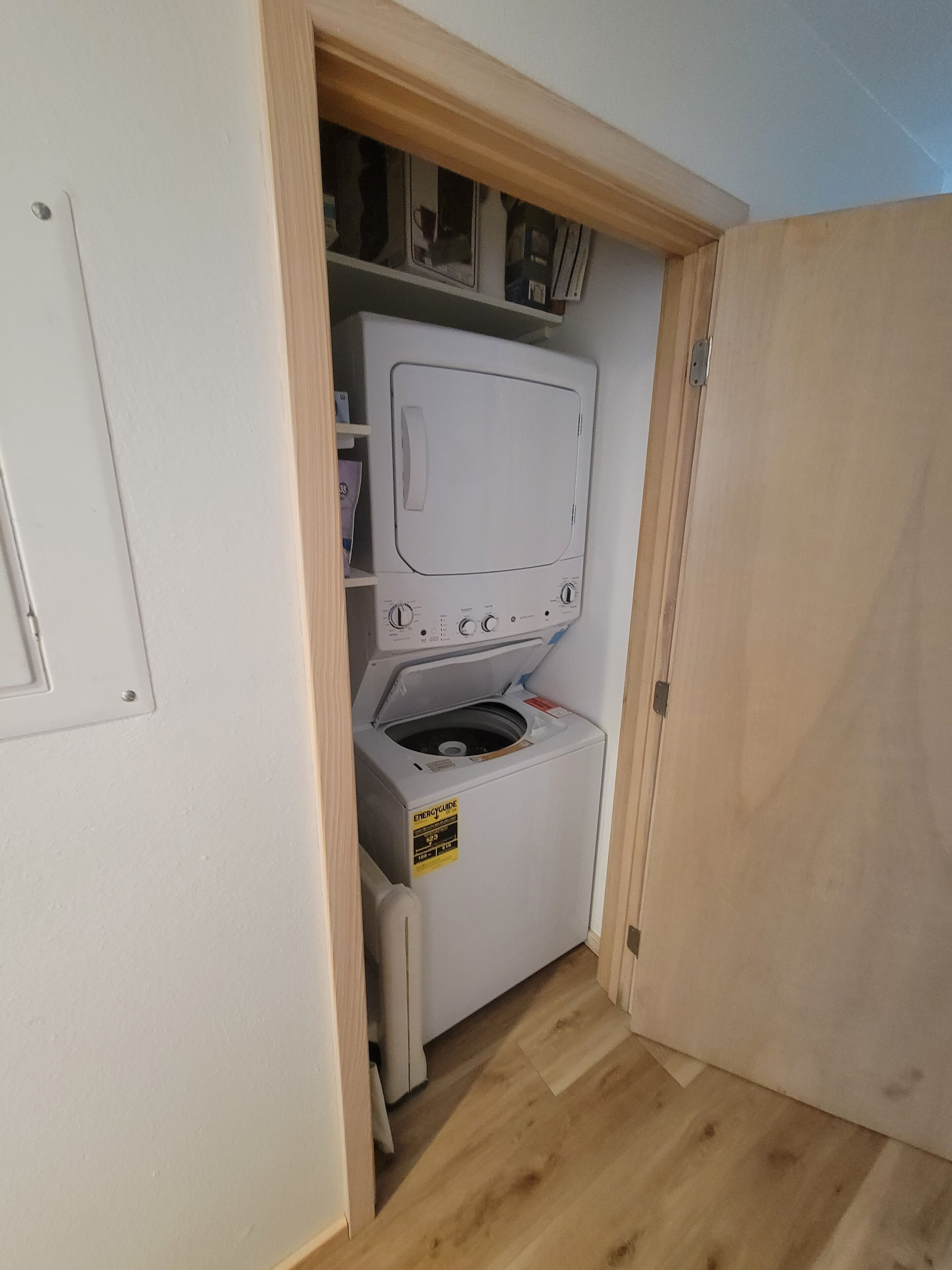 A compact laundry room with a front-loading washer and dryer, wooden flooring, and a small shelving unit above the appliances.