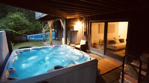 An outdoor hot tub area with a wooden deck, a glass door leading to a living room, and a pool in the background. - Secluded Getaway: Bear Mt, Cold Spring, Beacon, West Point!   (Garrison)