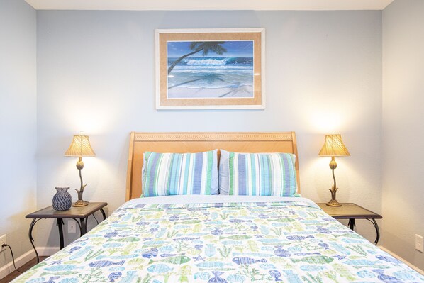 A bedroom with a bed, striped pillows, two bedside tables with lamps, a framed picture of a beach scene, and a patterned bedspread.