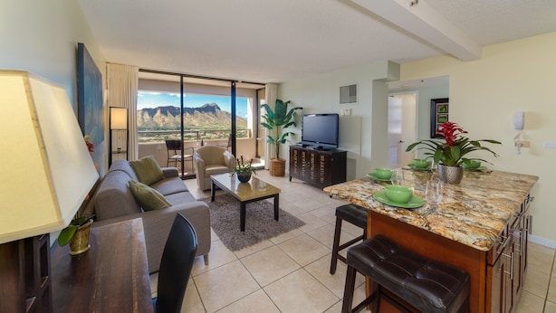 A modern living room with a sofa, coffee table, and TV, featuring a large window with a mountain view. - Amazing views of Diamond Head, Waikiki Beach, and the Pacific Ocean! (Honolulu)
