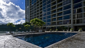 A swimming pool with lounge chairs and palm trees, surrounded by a modern building with multiple balconies. - Amazing views of Diamond Head, Waikiki Beach, and the Pacific Ocean! (Honolulu)
