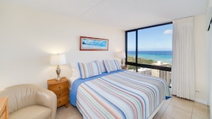 A bedroom with a large bed, a nightstand, a lamp, a chair, and a view of the ocean through a large window. - Amazing views of Diamond Head, Waikiki Beach, and the Pacific Ocean! (Honolulu)