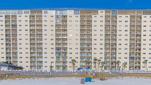 Un hotel de varios pisos con numerosas balconadas, una playa arenosa al frente y un cielo azul despejado.